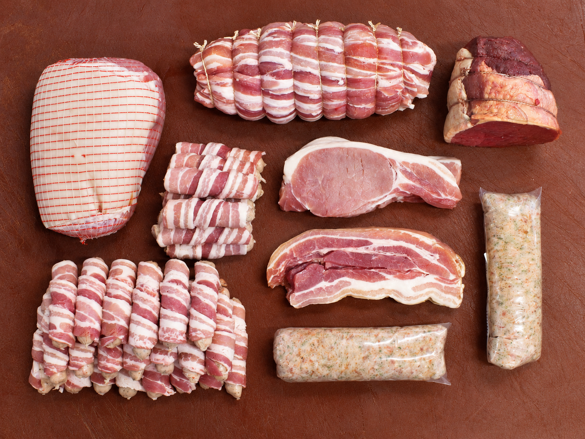 An assortment of raw cured meats and sausages arranged neatly on a dark brown wooden surface, including wrapped bacon rolls, sliced ham, a bone-in pork chop, and vacuum-sealed sausages.